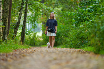 Obraz premium Girl walking the dog in the park forest. Rest concept. Woman walking with a dog on a leash in blue dress and with shoes.