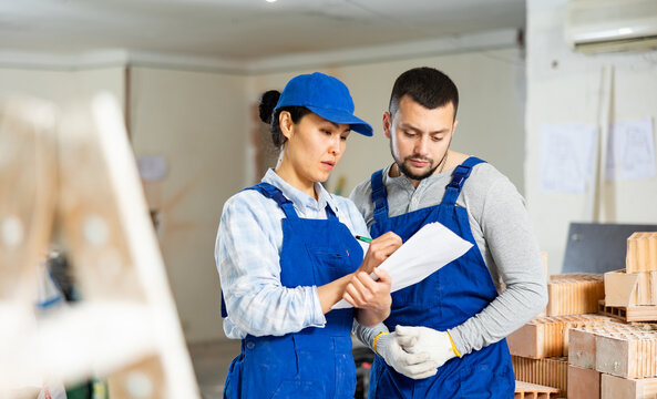 Focused skilled female and male builders in blue overalls determining scope of work and making list of tasks in building under renovation..