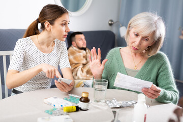 Women arguing about treatment of ill man at home. Senior woman reading instruction for medicine, her daughter-in-law searching information in internet with smartphone.