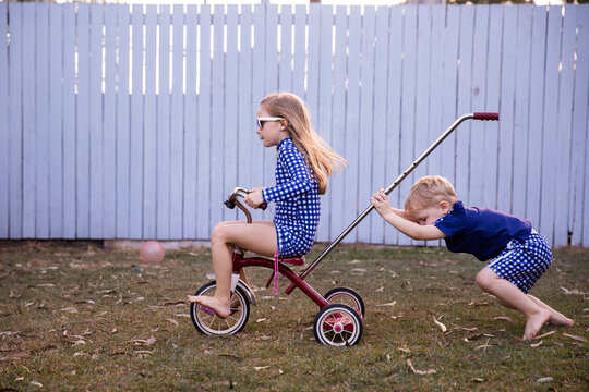 siblings playing on a trike in the back yard with a girl riding and a boy pushing