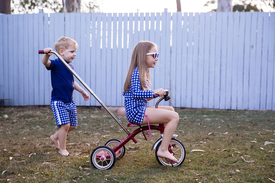 brother and sister playing on a trike in a backyard