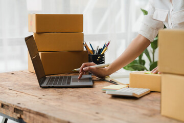 E-commerce Success: A woman's hand gracefully navigates a laptop keyboard, surrounded by stacks of...