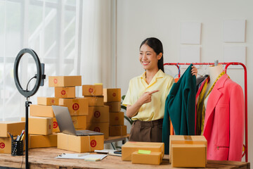 Online Fashion Entrepreneur: A smiling Asian woman presents clothing items while filming a video for her online shop, surrounded by packaging boxes and a laptop, showcasing the dynamic world of e-comm