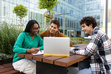 Happy multiracial university students studying and doing homework together outdoor college campus using laptop.