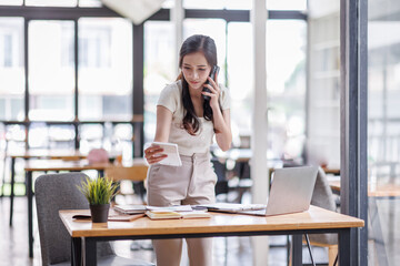 Smiling asian female hold cell at ear talk look at laptop screen. Busy young asian lady advisor...