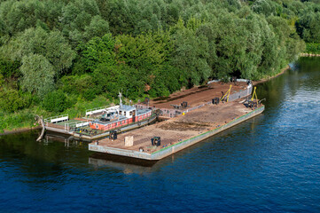 Docked barge and small boat by a green forested riverside.