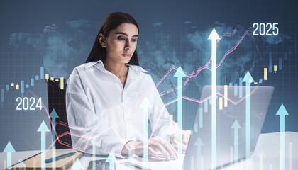 Attractive thoughtful young woman sitting at desk with laptop, supplies and growing business chart grid hologram and map on blurry background. Financial growth and fintech concept. Double exposure.