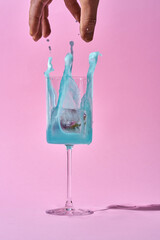 A splash of blue liquid erupts from a wine glass against a soft pink background. The liquid is frozen in mid-air, creating a dynamic and eye-catching composition.