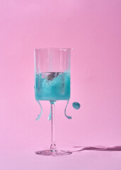 A splash of blue liquid erupts from a wine glass against a soft pink background. The liquid is frozen in mid-air, creating a dynamic and eye-catching composition.