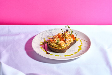 A gourmet open-faced sandwich on a white plate is adorned with grilled shrimp, a vibrant tomato salsa, and a creamy hummus spread.