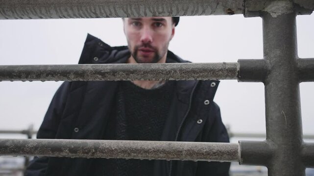 Man looking at camera through frozen metal fence in winter day outdoors. Portrait of handsome bearded man in warm clothes standing on city street alone, exhaling stream of air from mouth, stalker