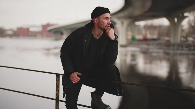 Carefree man in black jacket and hat sitting on rails of city embankment. Melancholy in autumn day, hipster in stylish casual clothes resting on quay alone, cool guy enjoying loneliness, street style