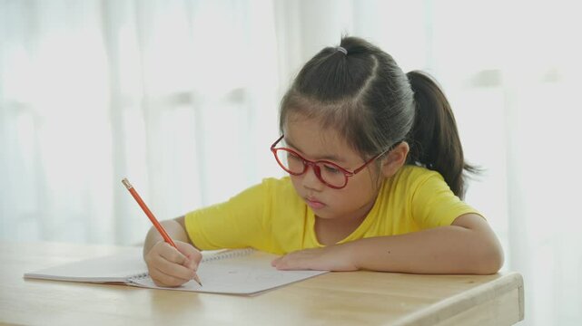Asian young girl is sitting at a table with a pencil and a notebook. She is wearing glasses and a yellow shirt. She is writing in her notebook, possibly a school assignment