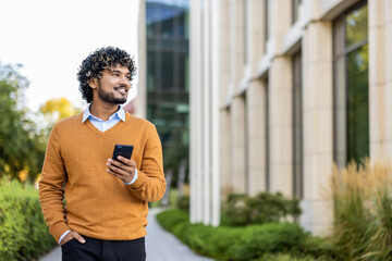 Hispanic businessman in orange sweater holding phone while walking outside office building. Smiling...