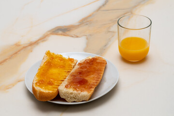 a couple of bread toasts with butter and strawberry and apricot jam on a white plate with a glass of orange juice on a white marbled table