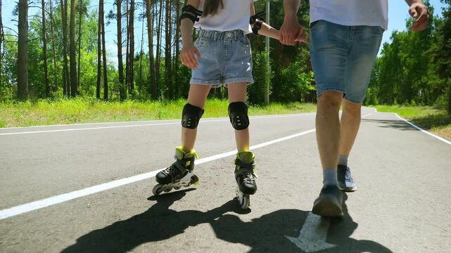 Young father walking towards camera on street and supporting little daughter while she learning how to roller skate