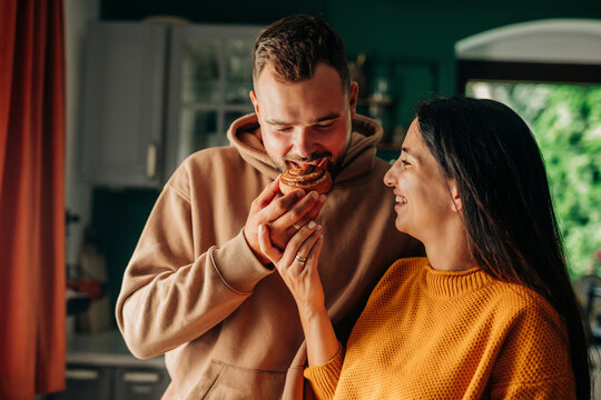 Young pregnant woman feeding cinnamon bun to husband in domestic kitchen