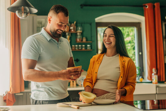Happy pregnant woman with husband holding muskmelon in kitchen at home