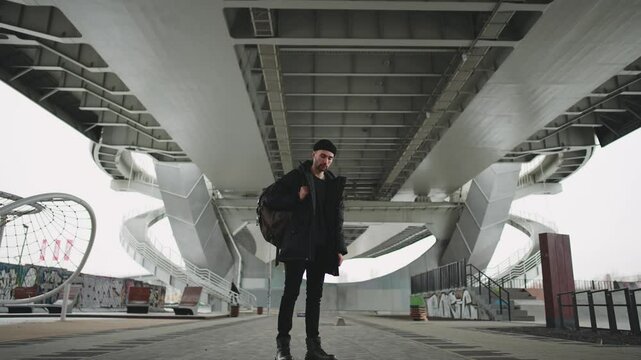 Young man in modern urbanistic recreation area for city dwellers in outskirts. Handsome hipster looking at camera, zooming shot, slow motion, contemporary lifestyle in town, infrastructure for sports