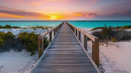 Obraz premium A long wooden boardwalk with a view extending to a white sandy beach and turquoise ocean at sunset. The sides of the boardwalk are adorned with occasional shrubs, enhancing the natural beauty.