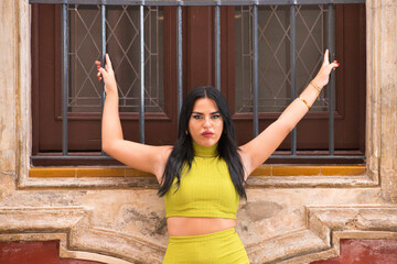 Young Spanish woman, brunette and beautiful in green dress leaning her back, hands and body against a red wall, the woman clings to the iron grille of a window grille of a stately home.