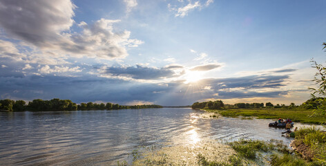 A river flows under a bright, cloudy sky with sunbeams shining through. People enjoy a boat ride on...