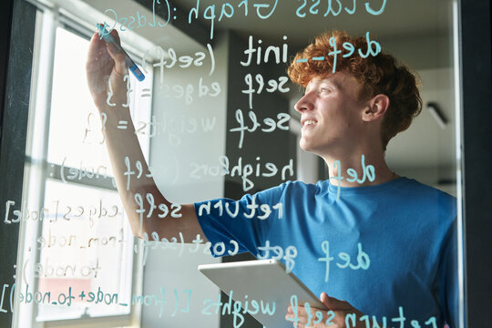 Side view of red haired man writing computer programming code on glass wall during IT class in school