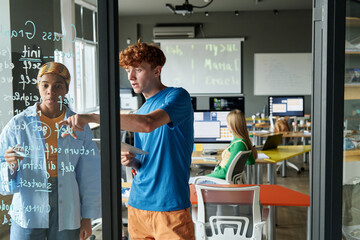 Two young people writing formulas on glass wall and collaborating on IT project together during...