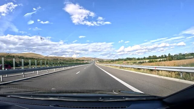 Driving on highway in Romania, surrounded by beautiful scenery with yellow hills and trees. Bright blue sky with white clouds overhead creates a serene atmosphere.