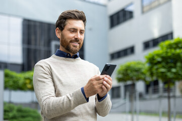 Confident businessman uses smartphone outside modern office building. Wearing casual attire, he checks messages or emails, showcasing connectivity and professional lifestyle in urban environment.