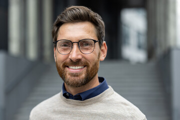 Portrait of confident man with glasses smiling warmly, conveying approachability and professionalism. Wearing stylish sweater, he stands in urban environment, suggesting success and positivity in