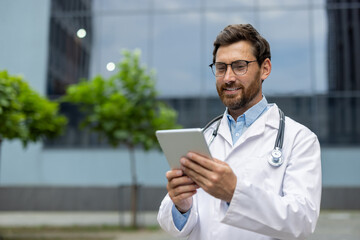 Doctor wearing white coat and stethoscope using tablet outside modern building. Smiling healthcare professional demonstrating confidence, embracing digital technology for patient care