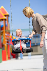 Mother pushing her infant baby boy child on a swing on playground outdoors