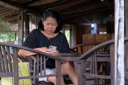Javanese woman sitting in her terrace of old wooden traditional house, reading messages on her smartphone