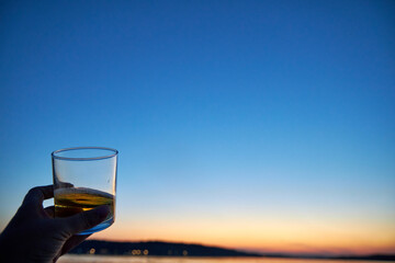 A glass with a drink in a woman's hand close-up against the background of a pink sunset on the water