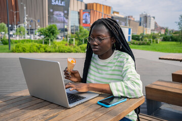 African american woman in glasses enjoys ice cream cone while working on laptop at outdoor table in...