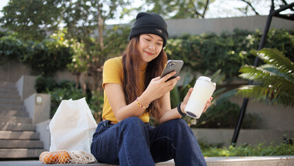 Gen Z eco go green woman asia sit happy relax at tree public park city life enjoy typing mobile phone drink coffee bottle use reuse tote bag care save the earth planet net zero waste clean future hope