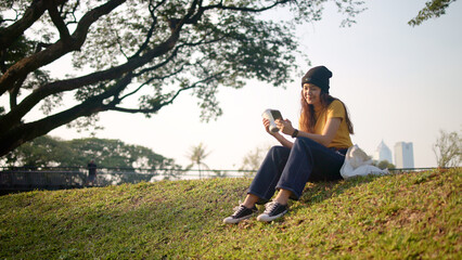 Gen Z eco go green woman asia sit happy relax at tree public park city life enjoy typing mobile...