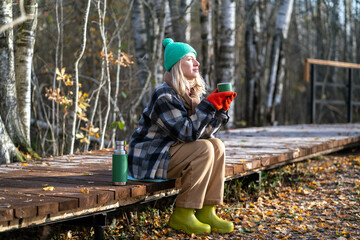 Satisfied middle aged woman spending tranquility time alone walking in autumn forest dreaming drinking hot thermos tea. Relaxed pleased female rest on eco path enjoys calm late fall park, stress free.