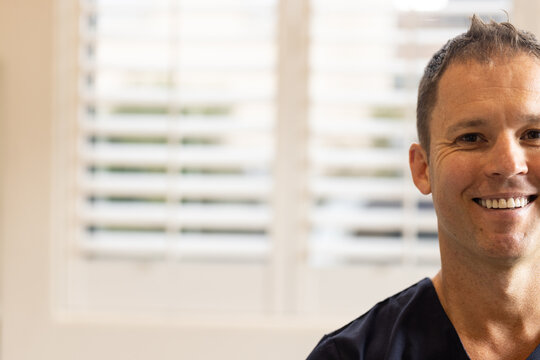 Smiling male doctor or healthcare worker in medical clinic wearing scrubs, standing near window blin