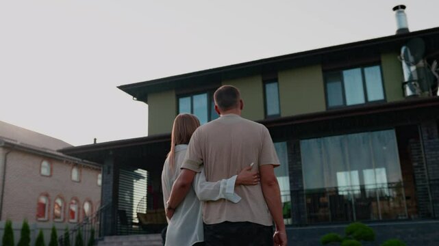 Back view of couple man and woman standing in front of huge house. Buying a house. Young just-married couple standing at yard of house, hugging and observing second floor