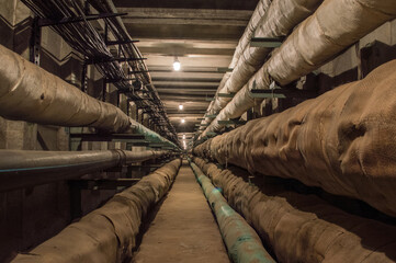 Underground concrete utility tunnel with pipes and wires lit by tungsten bulbs.. This tunnel gives...