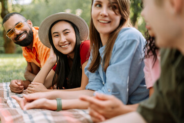 Group of smiling multiracial friends resting on blanket on green grass in park, relaxing
