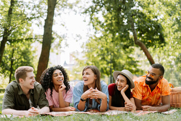 Portrait of happy multiracial friends, men and women relaxing, lying on blanket, having picnic