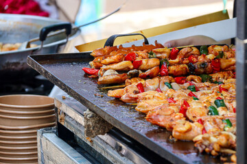 Close-up of sizzling grilled squid and sausages, garnished with vibrant red and green peppers on a hot griddle, showcasing the savory appeal of street food