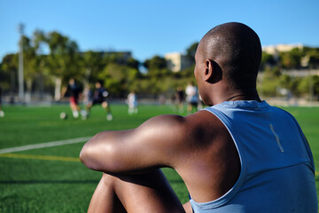 Rear back view athletic man soccer player wear blue athletic tank top and black shorts sitting on grassy sports football field, resting after game or workout. Take break, sports, lifestyle