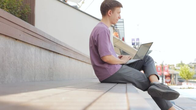 Smiling guy chatting social media at laptop outdoors. Young programmer working computer at street.