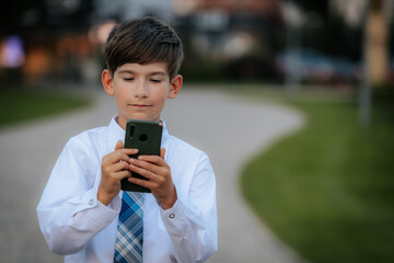 A young boy dressed in a white shirt and blue plaid tie is focused on his smartphone while walking in an urban park. The background shows a paved path and greenery