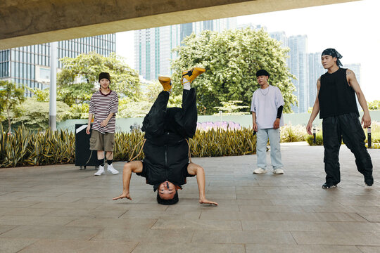 Group of young people practicing breakdance under modern city overpass with trees and buildings in background, breakdancer in center performing upside down move, wearing yellow shoes