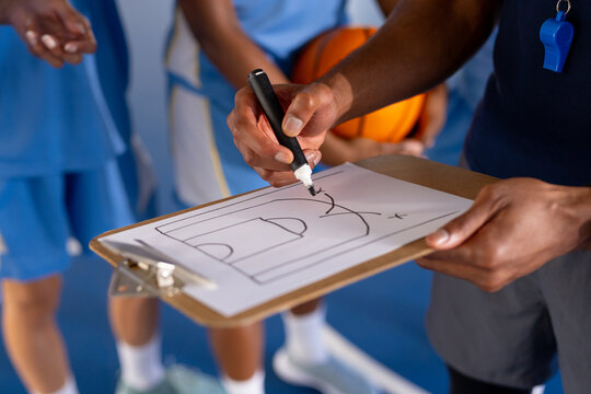 Male coach drawing basketball play on clipboard, players listening attentively in gym